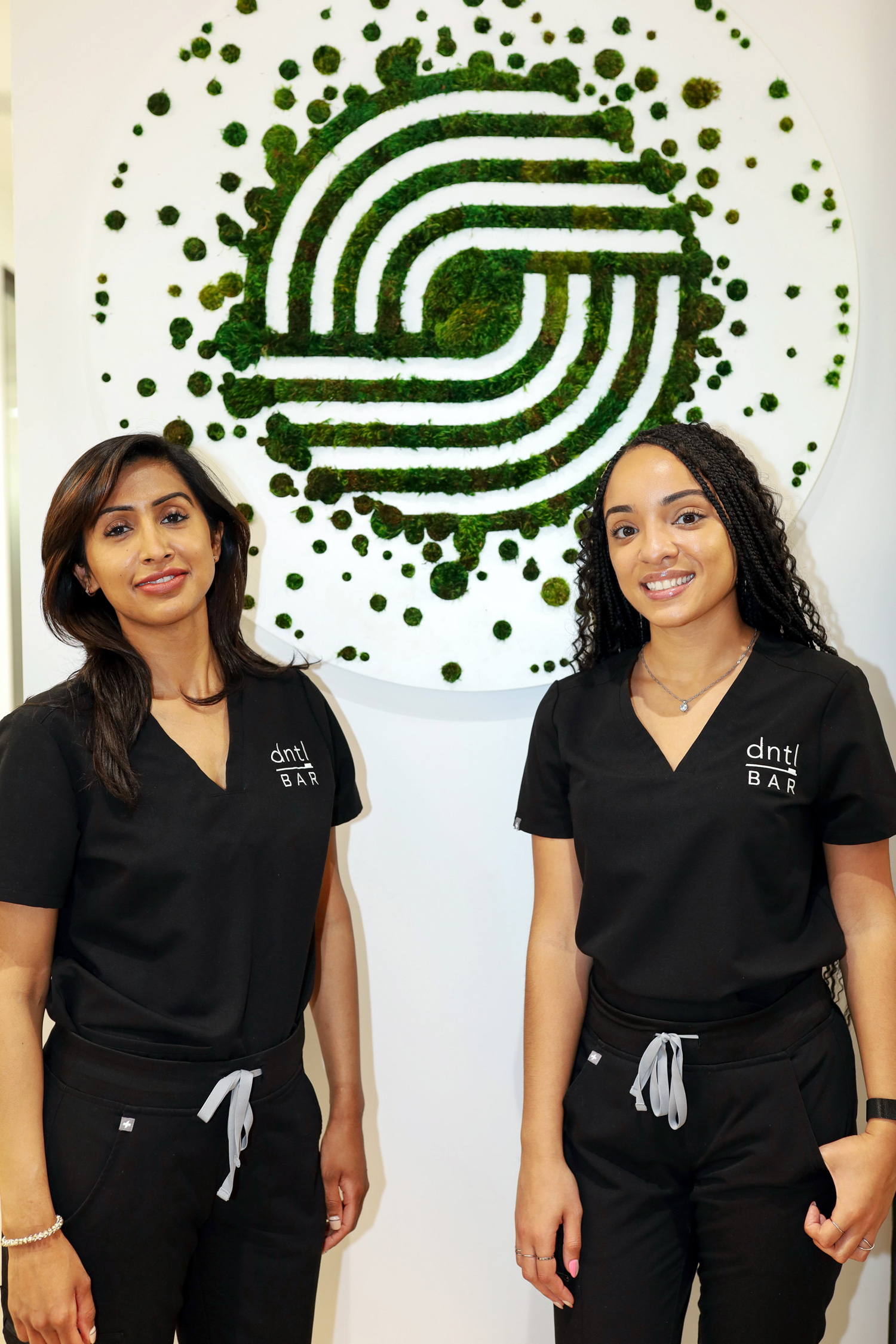 two dntl bar employees standing back to back smiling towards the camera against a moss artwork